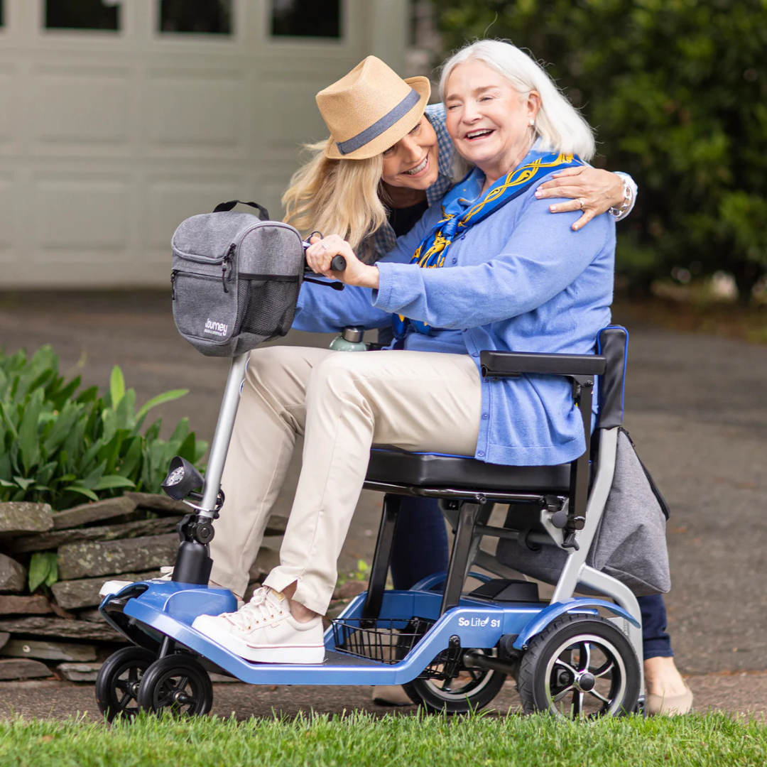 A joyful elderly woman rides a Journey So Lite® Folding Power Scooter, hugging a middle-aged woman in a hat. They smile and embrace in a garden with greenery and a garage door behind them. The scooters lightweight design and Electronic Anti-Tip Technology ensure her comfort and safety.