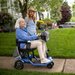 An elderly woman rides her Journey So Lite® Folding Power Scooter, its lightweight design aiding her navigation on the scenic residential street lined with lush grass and flowers. Smiling, she chats with a younger woman walking beside her, confident in the scooters electronic anti-tip stability.