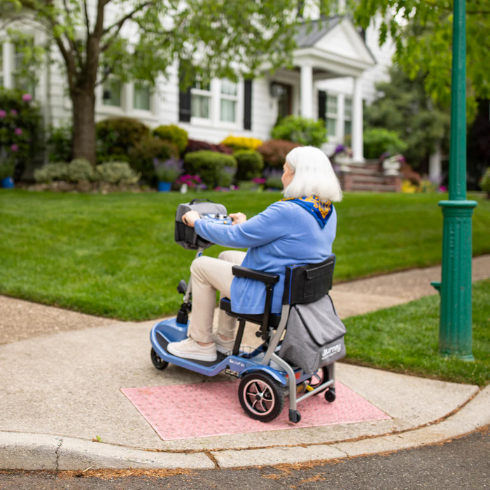 An older woman with white hair rides a Journey So Lite® Folding Power Scooter on the sidewalk. Wearing a blue sweater and beige pants, she enjoys added stability from the electronic anti-tip technology, with a green lawn and flower-adorned white house in the background.