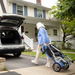 A woman in a blue sweater is folding her Journey So Lite® Folding Power Scooter and placing it into a car trunk. In the background, theres a white house with blue shutters amid green trees and grass.