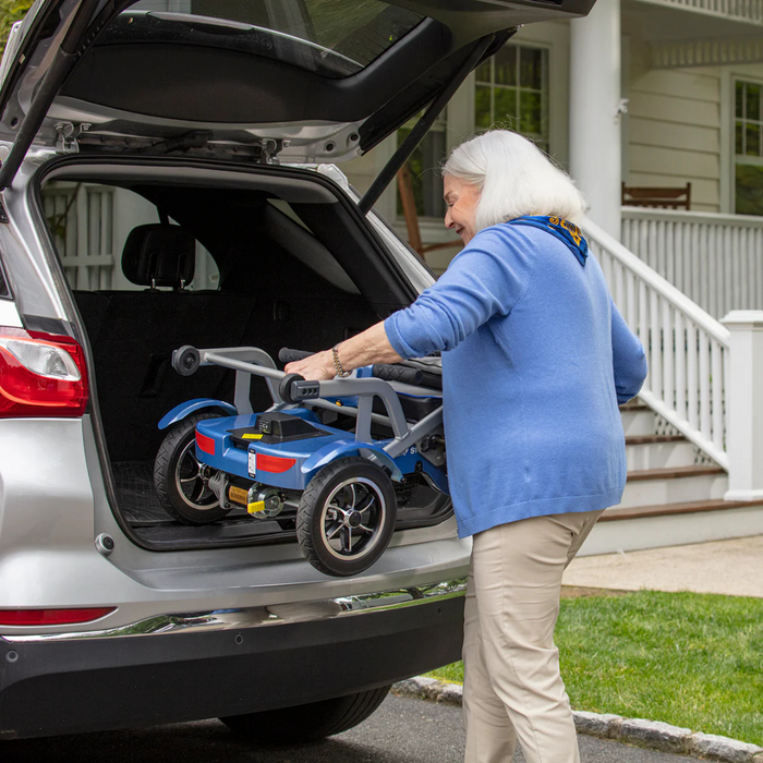 A white-haired woman in a blue sweater easily places a folded Journey So Lite® Folding Power Scooter—famous for its lightweight design and electronic anti-tip tech—into a silver SUVs trunk, with a charming white house and porch elegantly featured in the background.