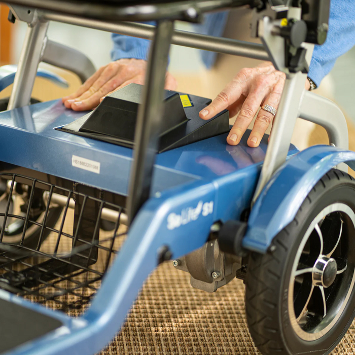 A person adjusts a part on a blue Journey So Lite® Folding Power Scooter. Their hands are near the rear wheel, and a basket is visible beneath. Indoors on carpet, this lightweight scooter offers advanced electronic anti-tip technology for safety and stability.