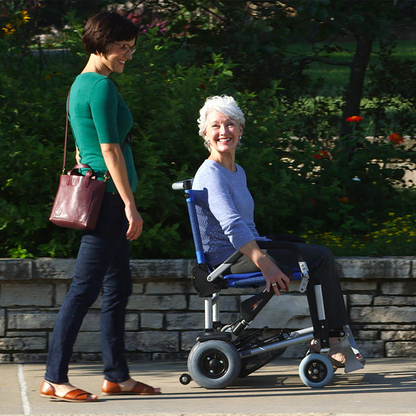 A woman with short gray hair is seated in a Journey Zinger® Folding Powerchair (FAA Approved, 47 lbs), smiling as she moves along a path. Beside her walks another woman with short dark hair, also smiling. They are outdoors, surrounded by lush greenery and flowers.