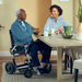 An elderly man in a Journey Zoomer® Portable Powerchair and a woman sit at a warmly lit dining table, chatting and smiling. The table holds mugs, a green vase, and a fruit bowl against light-colored walls, creating an inviting scene.