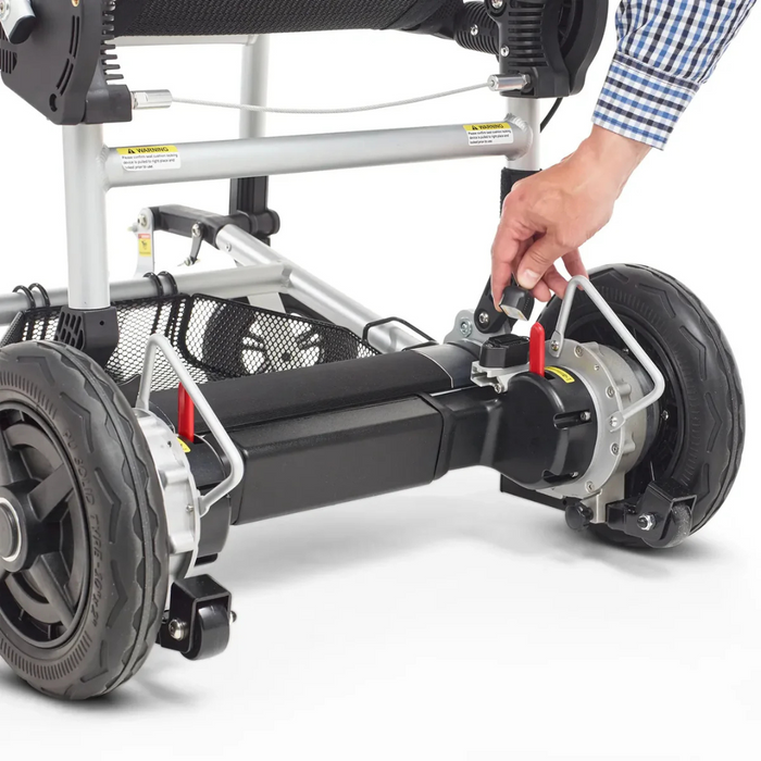 Close-up of a person in a blue and white checkered shirt adjusting the brake lever on the rear wheels of the Journey Zoomer® Portable Travel Powerchair. With a metal frame and large black wheels, this innovative design ensures a compact turning radius.