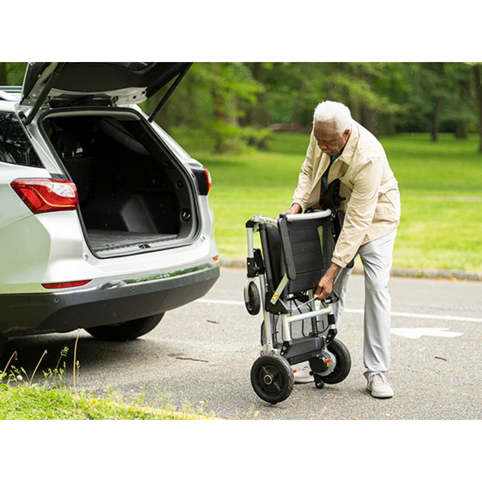 An elderly man folds the Journey Zoomer® Portable Travel Powerchair—FAA Approved—next to the open trunk of a white SUV in a park setting with lush green trees and grass.