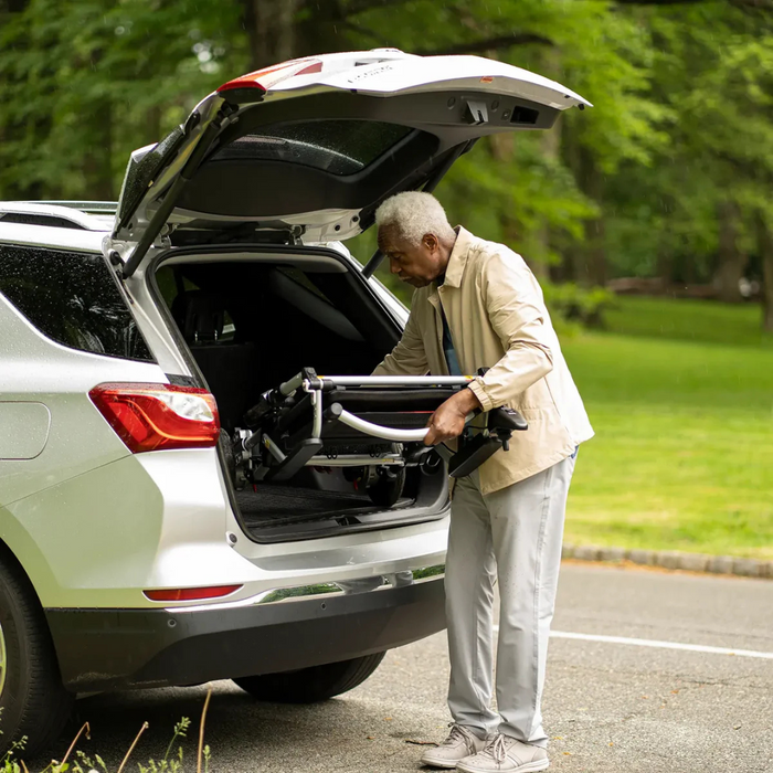 An older adult in a beige jacket and gray pants lifts the Journey Zoomer® Portable Travel Powerchair into the back of a silver SUV, against a backdrop of a grassy park.