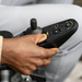 The image shows a hand using the joystick on the innovative Journey Zoomer® Portable Travel Powerchair, FAA Approved. The control panel displays various settings, and the hands position indicates movement or adjustment, highlighting the chairs compact turning radius.