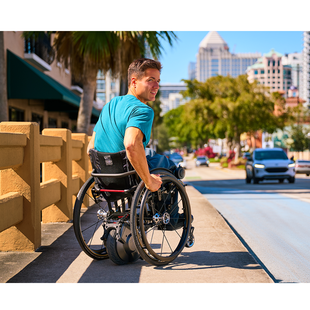A man in a wheelchair with a Klaxon® Twist Mobility Power-Assist Hub | Wheelchair Power Assist smiles while using a sidewalk ramp in a sunny city with cars, palm trees, and tall buildings in the background.