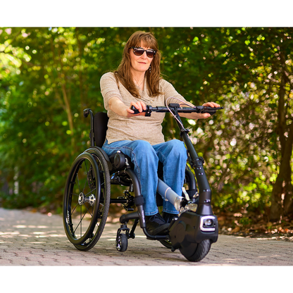 A woman wearing sunglasses rides a manual wheelchair with a Klaxon® Twist Mobility Power-Assist Hub | Wheelchair Power Assist attached, along a sunny, tree-lined path.