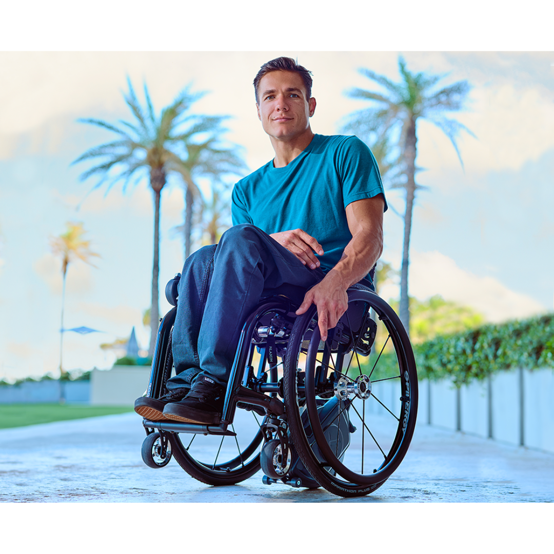 A man in a wheelchair with a Klaxon® Twist Mobility Power-Assist Hub | Wheelchair Power Assist smiles outdoors on a sunny day, with palm trees and blue sky behind him. He wears a blue shirt and dark pants.