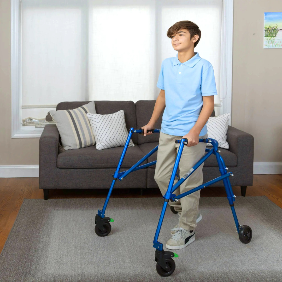 A boy in a light blue shirt and khaki pants uses a Circle Specialty Klip Pediatric Posterior Walker in a living room with a gray sofa, striped pillows, and a large window in the background.