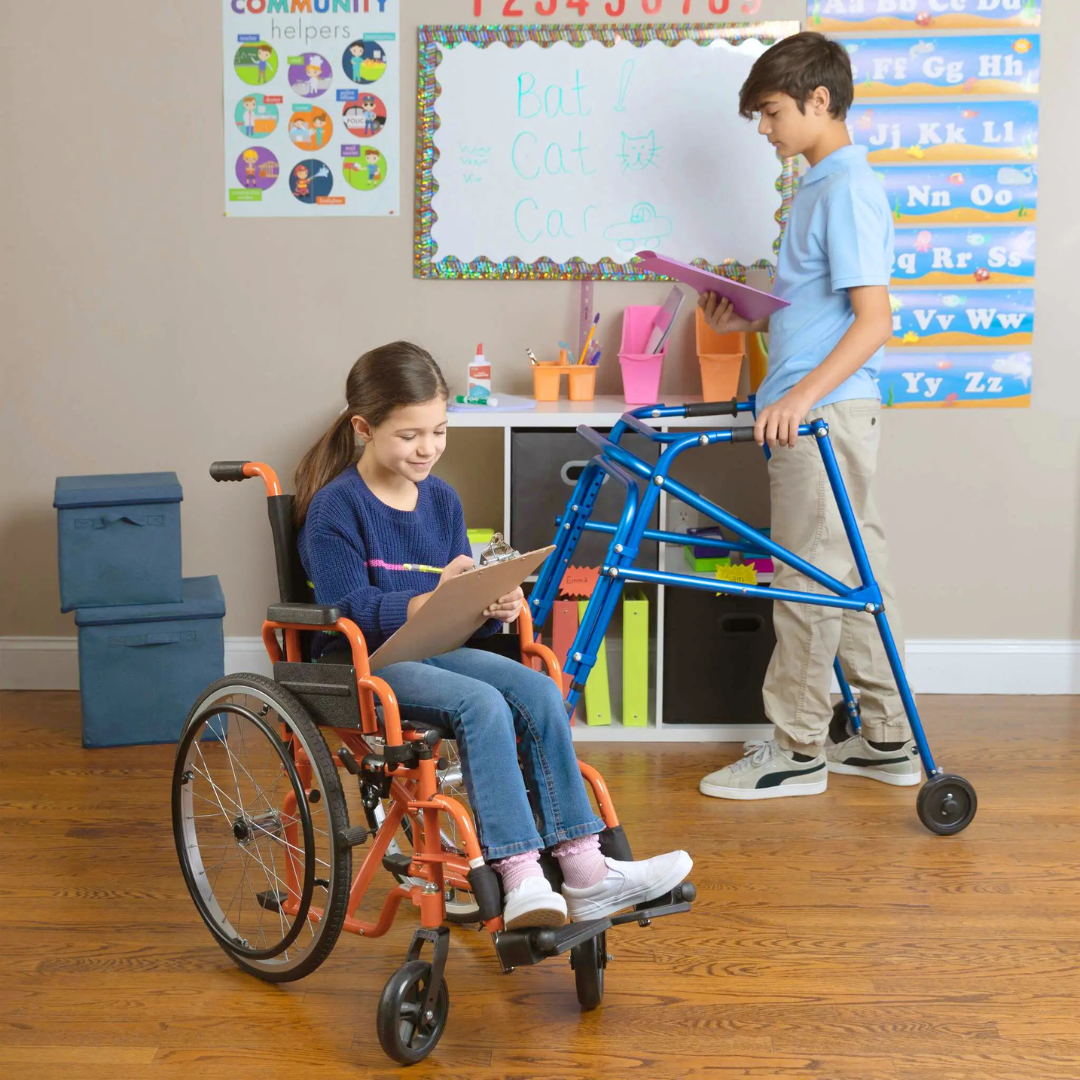 A girl in a wheelchair and a boy using the Circle Specialty Klip Pediatric Posterior Walker write on clipboards in a classroom, with educational posters and storage bins lining the shelves behind them.