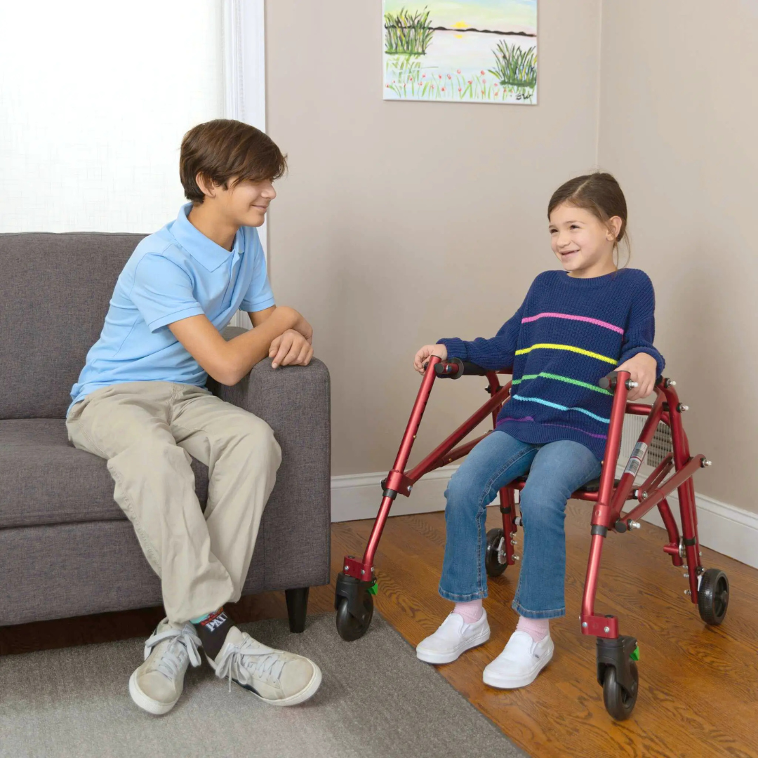 A young girl in a red pediatric wheelchair smiles at a boy on a gray couch. The Circle Specialty Klip Pediatric Posterior Walker stands nearby on the wood floor under a colorful painting, enhancing the welcoming indoor scene.