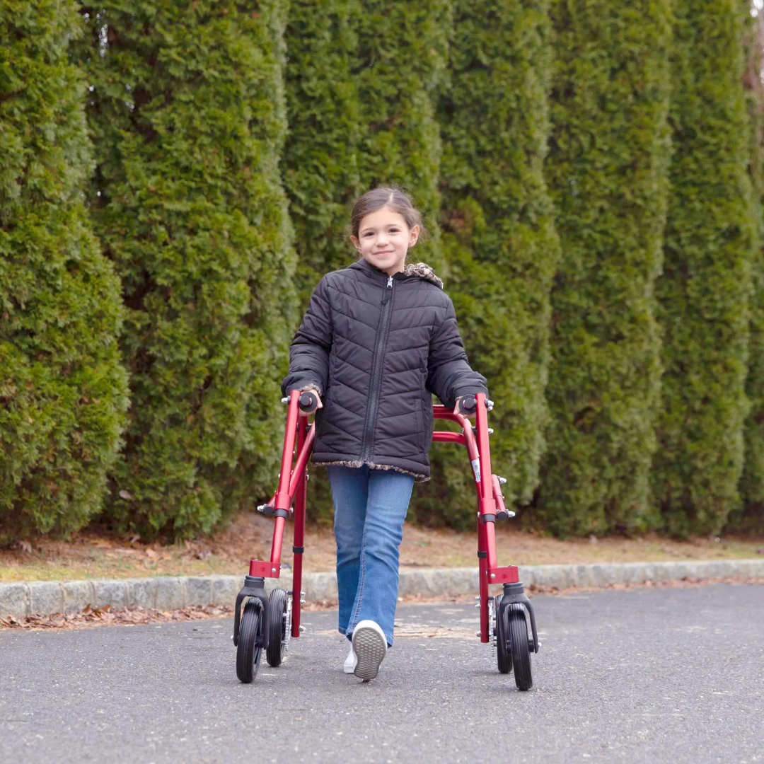 A young girl in a black jacket and jeans smiles as she walks outdoors along a path with tall green bushes, using the Circle Specialty Klip Pediatric Posterior Walker to support her mobility.