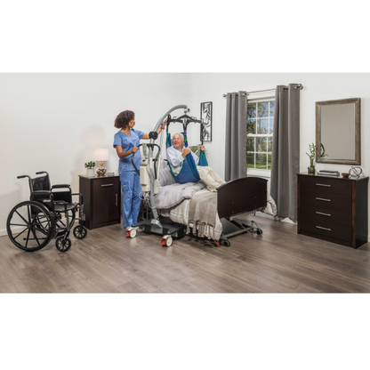 A nurse helps an elderly patient in a hospital bed using the Drive Levantar Battery-Powered Patient Floor Lift, with a wheelchair nearby in a clean, well-lit room featuring wood floors, modern furniture, and gray curtains.