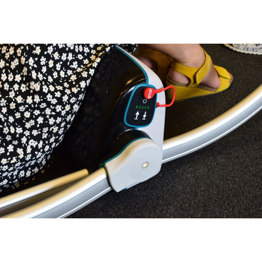 A person in yellow sandals and a black-and-white floral skirt sits beside the LiftUp Raizer II, their foot near its control panel with a red emergency stop button, indicator arrows, and the user manual visible nearby.