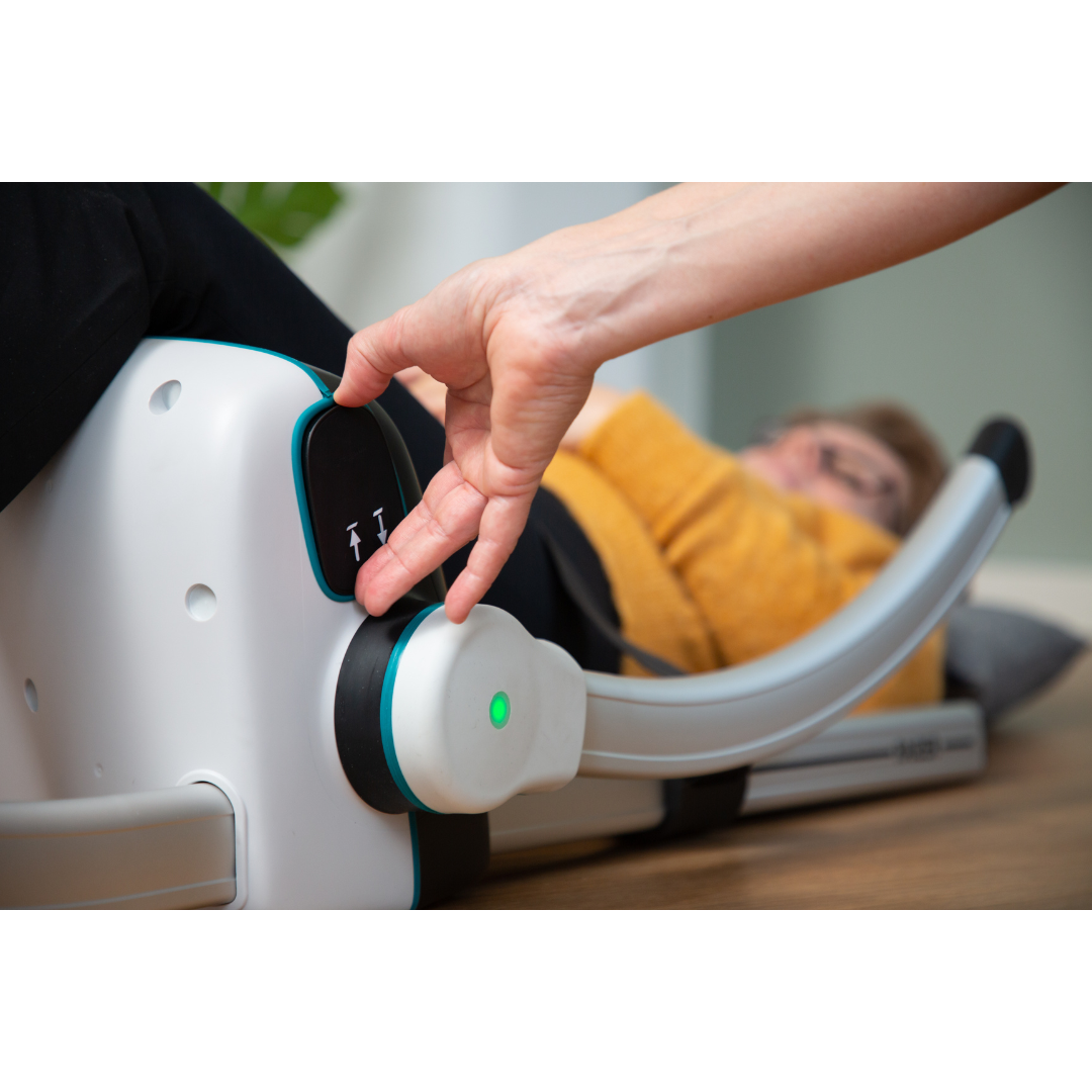 A person adjusts the control knob with arrow icons on the LiftUp Raizer II therapy device, which emits a glowing green light. The scene is indoors on a wooden floor, with a user manual nearby.