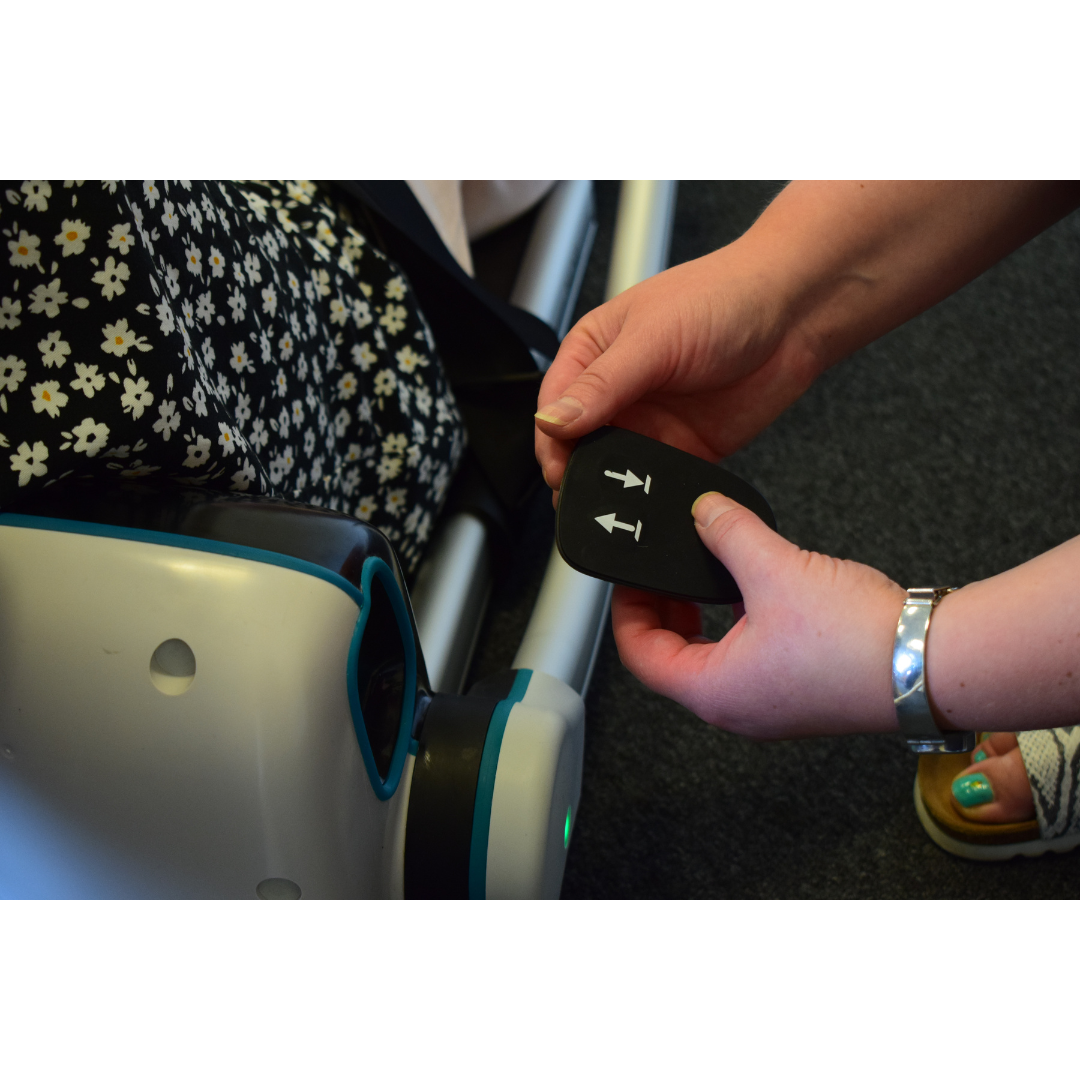 A person adjusts the LiftUp Raizer IIs black strap with white arrow symbols on a stroller, following the user manual. A floral-patterned bag hangs on the stroller, and they wear a silver bracelet and sandals.