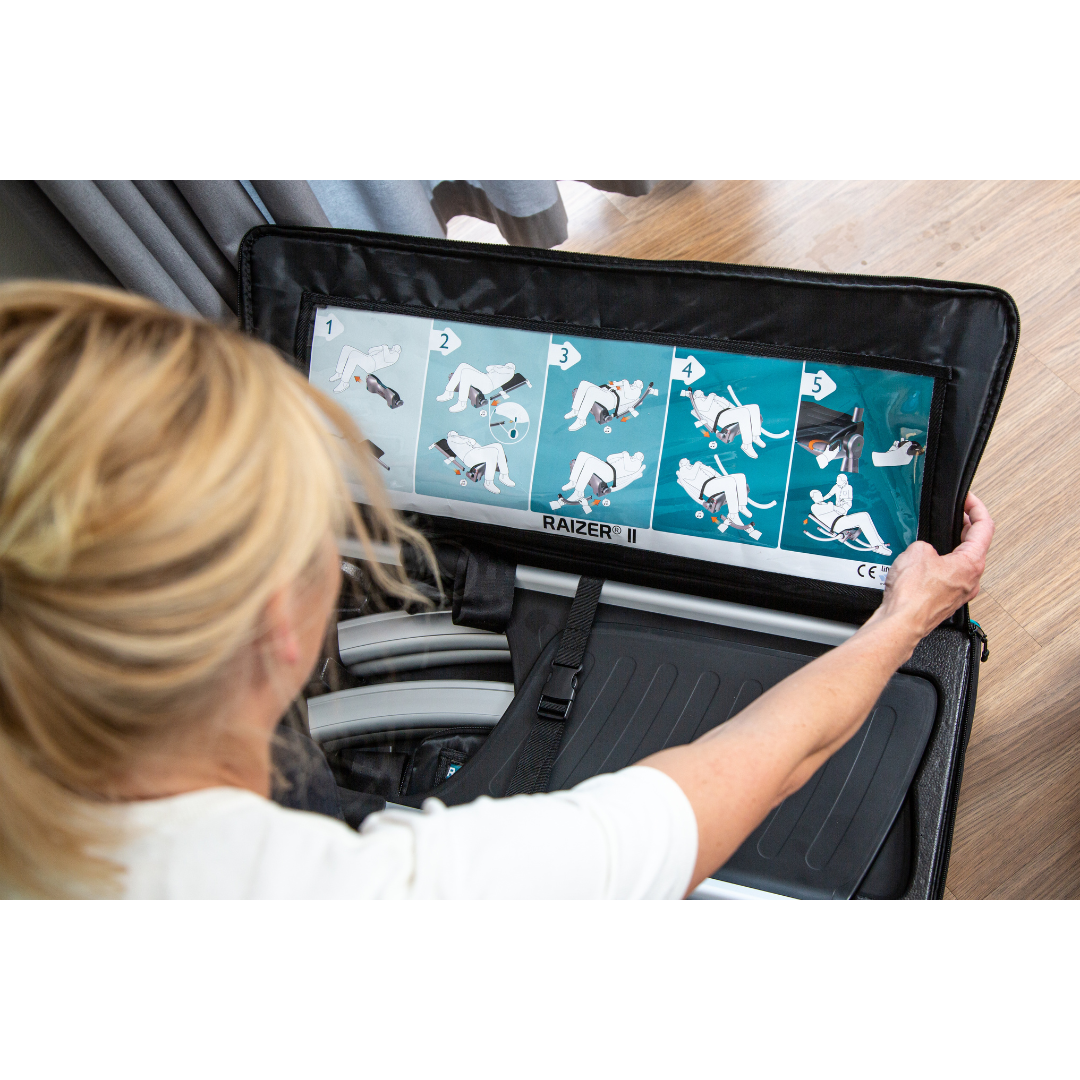 A woman reviews illustrated step-by-step instructions inside the lid of a black LiftUp Raizer II case, likely to guide her in using this medical lifting device. The partially open case sits on a wooden floor.