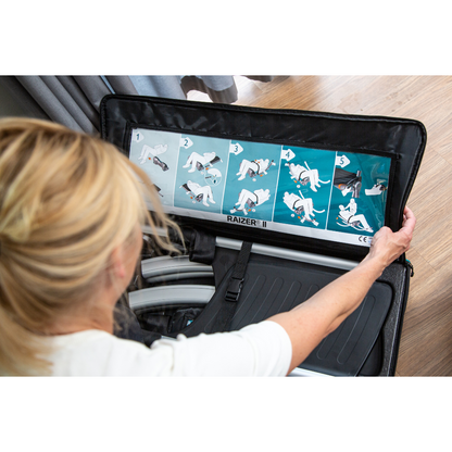 A woman reviews illustrated step-by-step instructions inside the lid of a black LiftUp Raizer II case, likely to guide her in using this medical lifting device. The partially open case sits on a wooden floor.