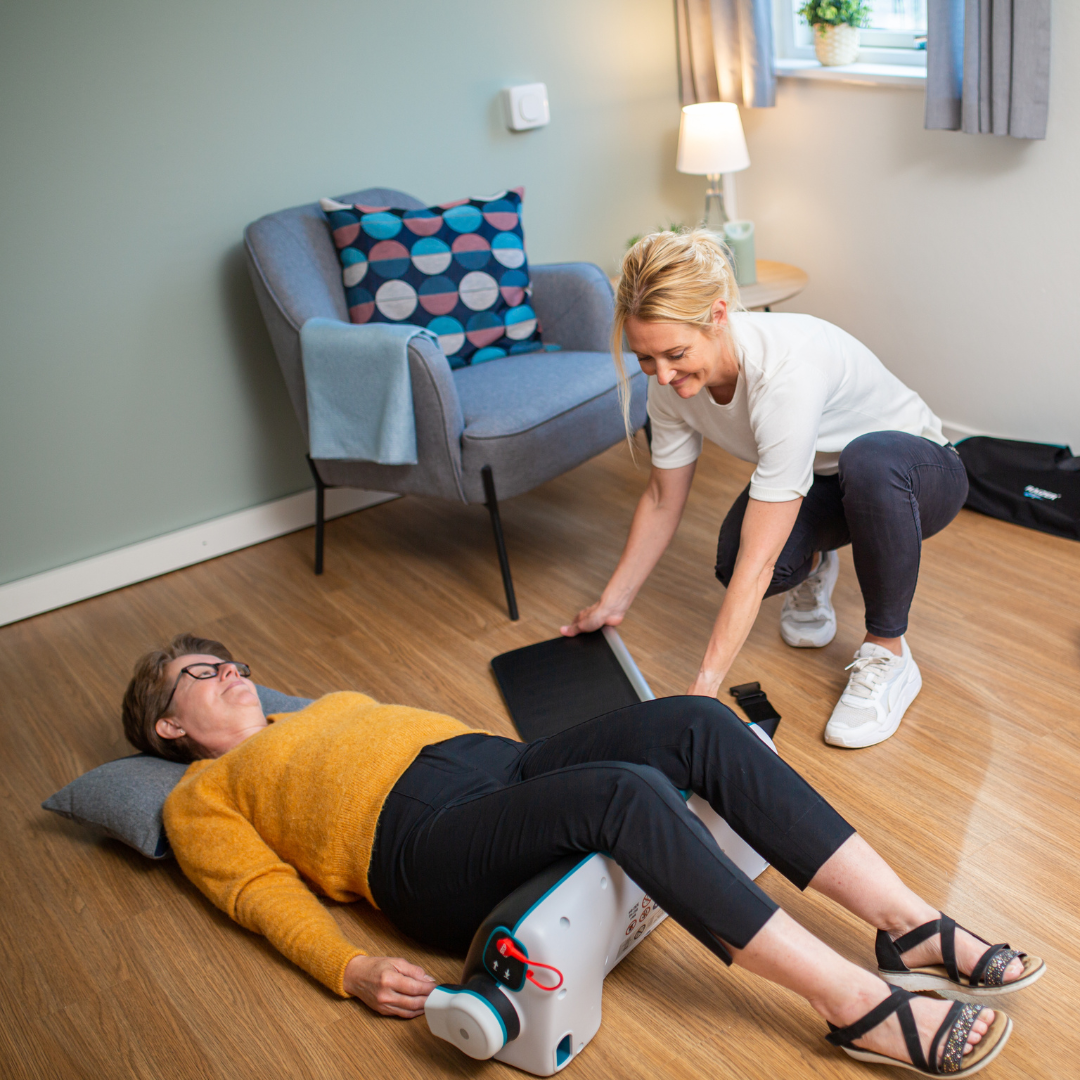 A woman assists another woman lying on the floor with her legs elevated on the LiftUp Raizer II, referring to the user manual in a cozy room with wood flooring, an armchair, and a lamp.