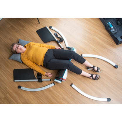 A woman in glasses, a yellow sweater, and black pants lies on a wooden floor with her head on a pillow, surrounded by LiftUp Raizer II metal and plastic parts as she references the user manual to assemble the equipment.