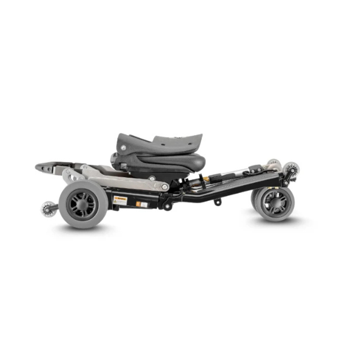 The FreeRider Luggie Standard Travel Scooter is shown fully folded, featuring grey wheels and a compact black frame, designed as a portable mobility aid for effortless transport and storage.