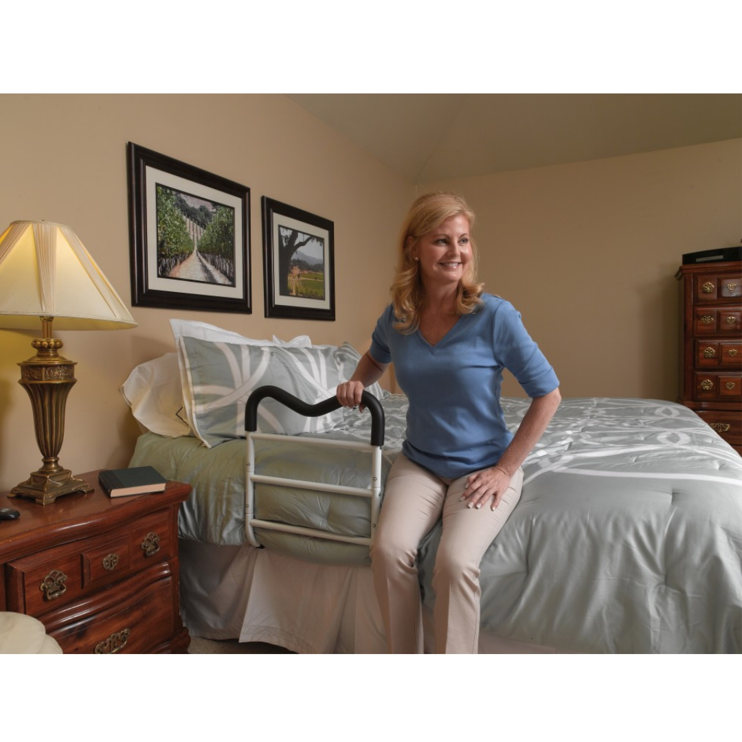 A woman sits on the edge of a bed, holding a Drive M-Rail Home Bed Assist Handle. The warmly lit room includes framed pictures, a lamp, and wooden furniture such as a nightstand and dresser.