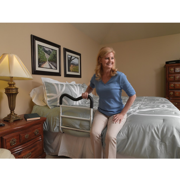 A woman sits on the edge of a bed, holding a Drive M-Rail Home Bed Assist Handle. The warmly lit room includes framed pictures, a lamp, and wooden furniture such as a nightstand and dresser.
