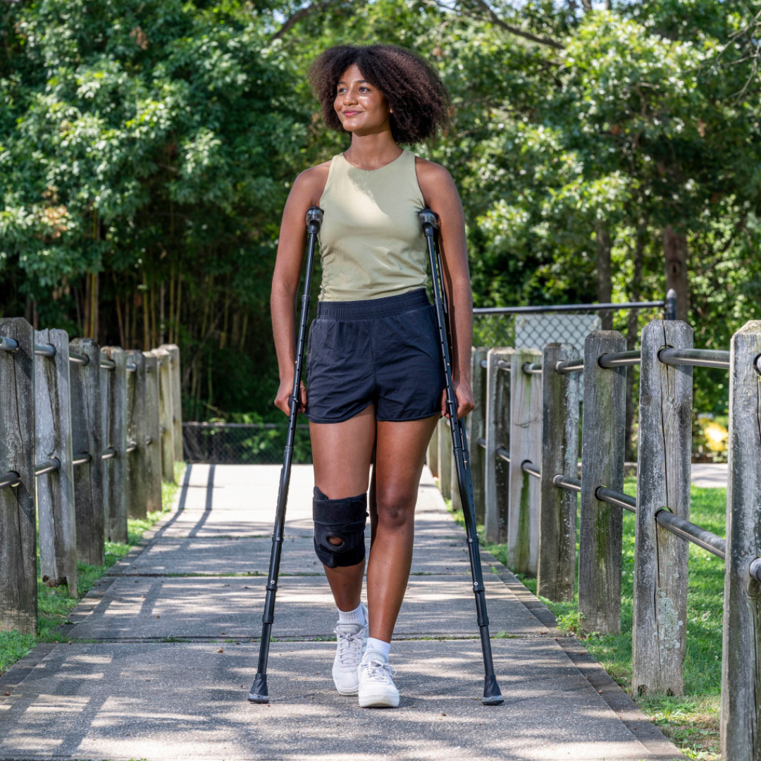 A woman with curly hair walks outside on a path using Drive Air-Crutches with Airgonomic™ Underarm Cushions. She wears a knee brace, shorts, a sleeveless top, and sneakers. Trees and a wooden fence line the path.