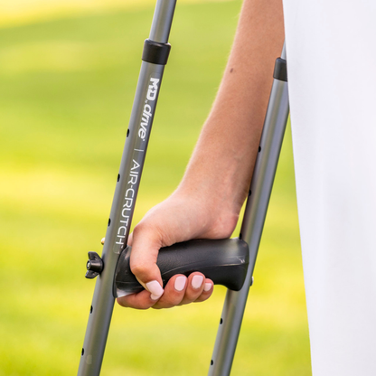 A person grips the handle of a gray Drive Air-Crutches with Airgonomic™ Underarm Cushions, underarm pads visible, against a blurred green outdoor background.