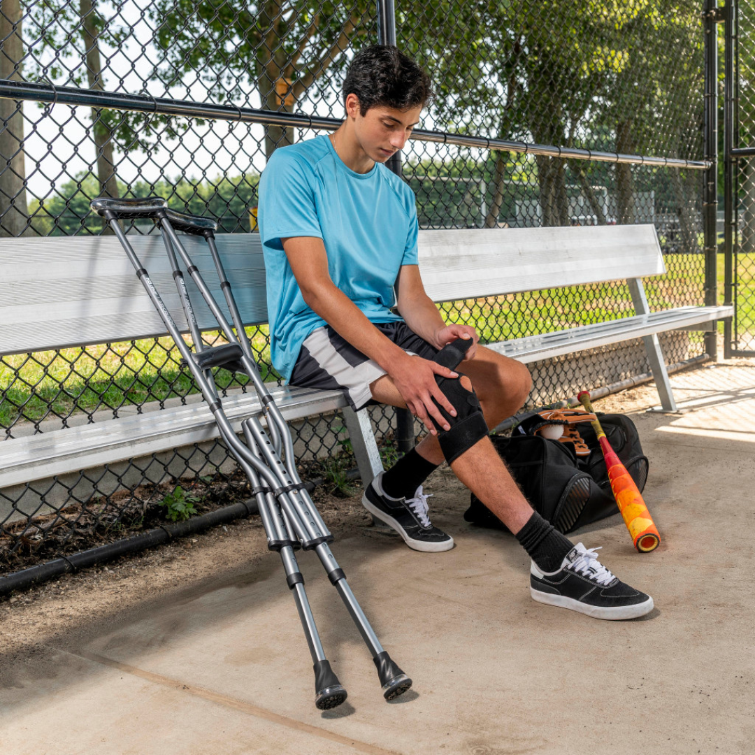 A teenage boy sits on a bench by a baseball field, his knee in a brace. Next to him are Drive Air-Crutches with Airgonomic™ Underarm Cushions, a baseball bat, and a sports bag. Trees and chain-link fencing are in the background.