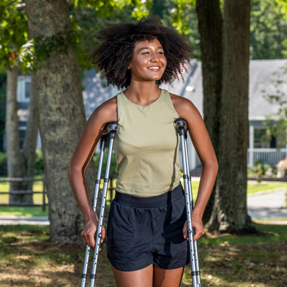 A young woman with natural hair smiles while walking outside in a sunny, leafy neighborhood using Drive Air-Crutches with Airgonomic™ Underarm Cushions. She wears a sleeveless top and shorts, with trees and houses in the background.
