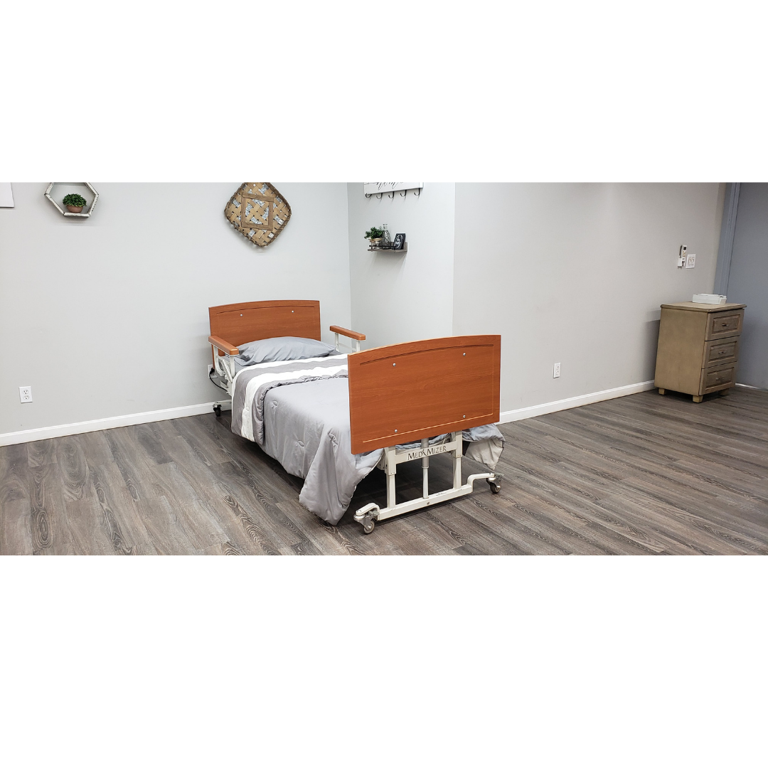 A Med-Mizer AllCare Full Electric Hi/Low Bed - Expandable Width with wooden head and footboards is in a clean room with gray walls, wood floors, minimal decor, and a small cabinet in the corner.