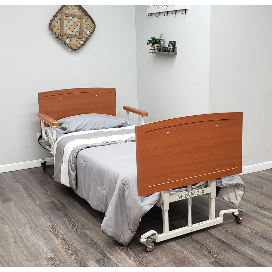 A Med-Mizer AllCare Full Electric Hi/Low Bed - Expandable Width with wood panel headboard and footboard, gray and white bedding sits on wood flooring in a light gray room. A wall shelf and decorative basket are mounted on the walls.