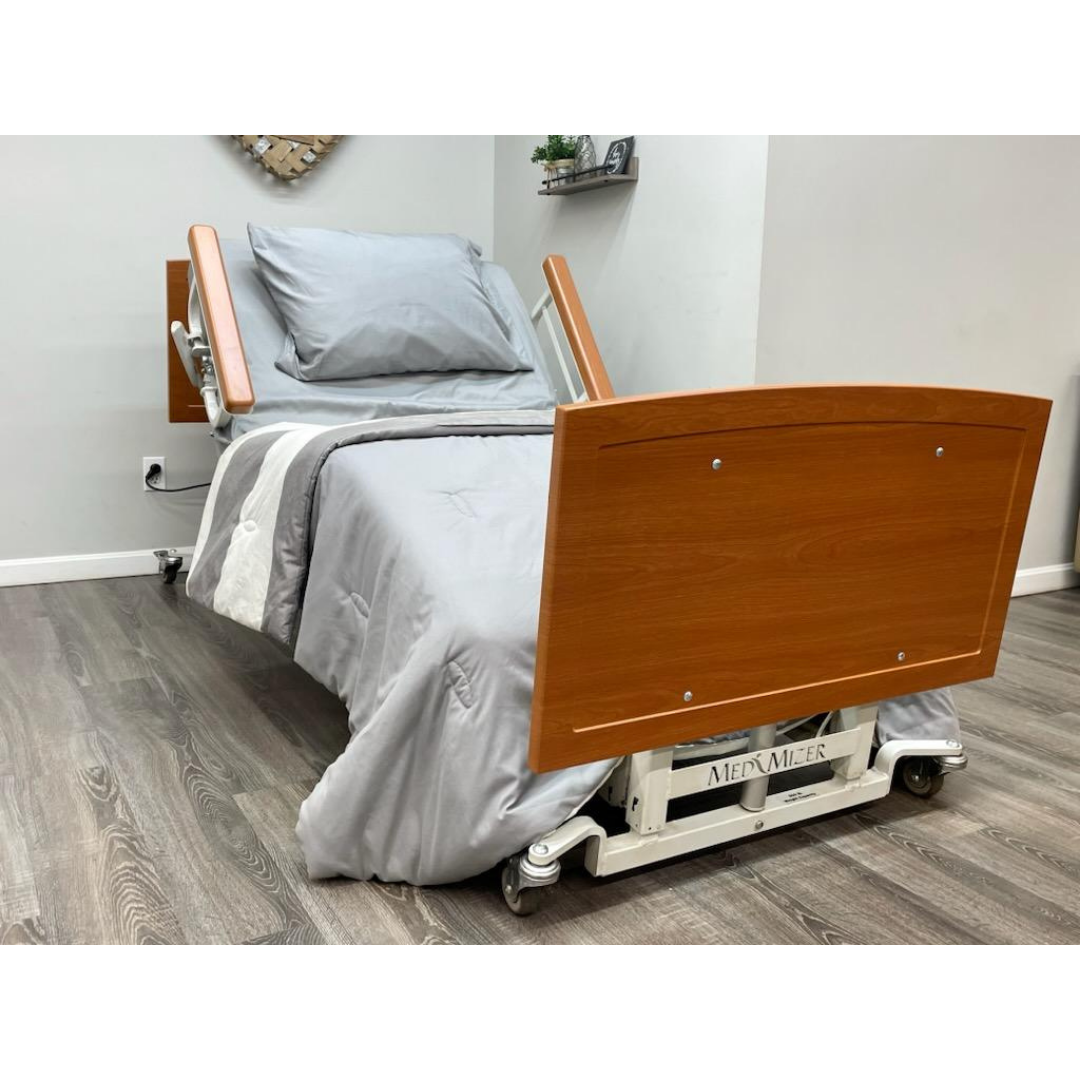 A Med-Mizer AllCare Full Electric Hi/Low Bed - Expandable Width with wood headboard and footboard stands in a clean room with gray bedding, adjustable side rails, wood flooring, and decorative shelves on the wall.