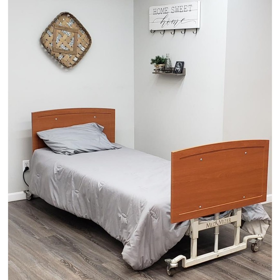 A Med-Mizer AllCare Full Electric Hi/Low Bed with light gray bedding is in a cozy room with wood flooring, decorated with a woven basket, “Home Sweet Home” sign, and a small shelf holding plants and decor.