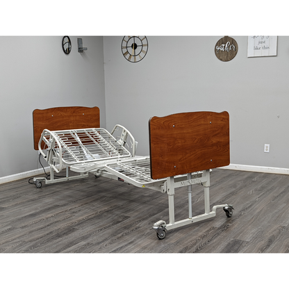 A Med-Mizer AllCare Full Electric Hi/Low Bed - Expandable Width with a wooden headboard and footboard sits in a room with gray walls, wood flooring, and wall decor such as clocks and framed quotes.