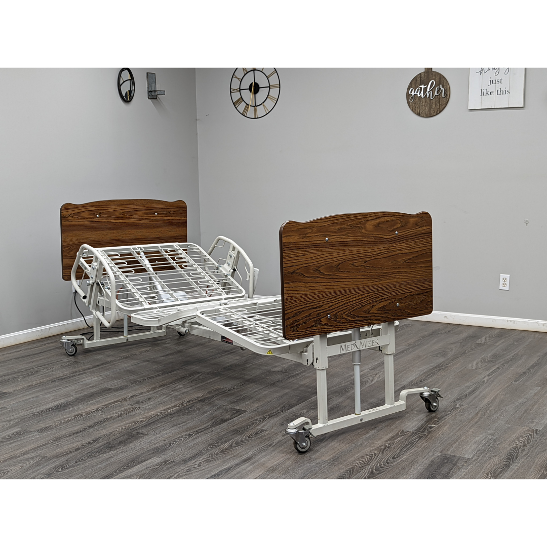 A Med-Mizer AllCare Full Electric Hi/Low Bed - Expandable Width with wooden headboard and footboard sits in a room with gray walls, wood flooring, and wall decor like clocks and a "gather" sign. The bed is empty and partially elevated.