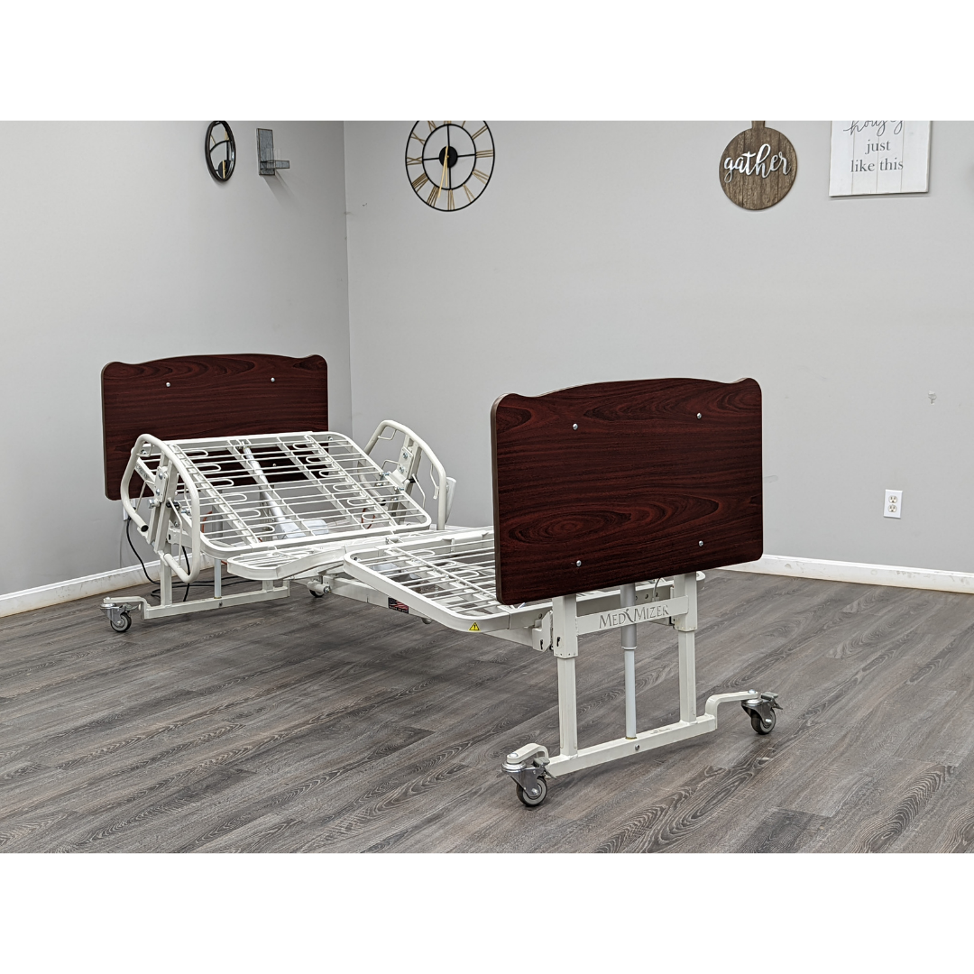 A Med-Mizer AllCare Full Electric Hi/Low Bed - Expandable Width with wooden headboard and footboard on wheels sits in a room with gray walls, wood floors, clocks, and inspirational wall decor.