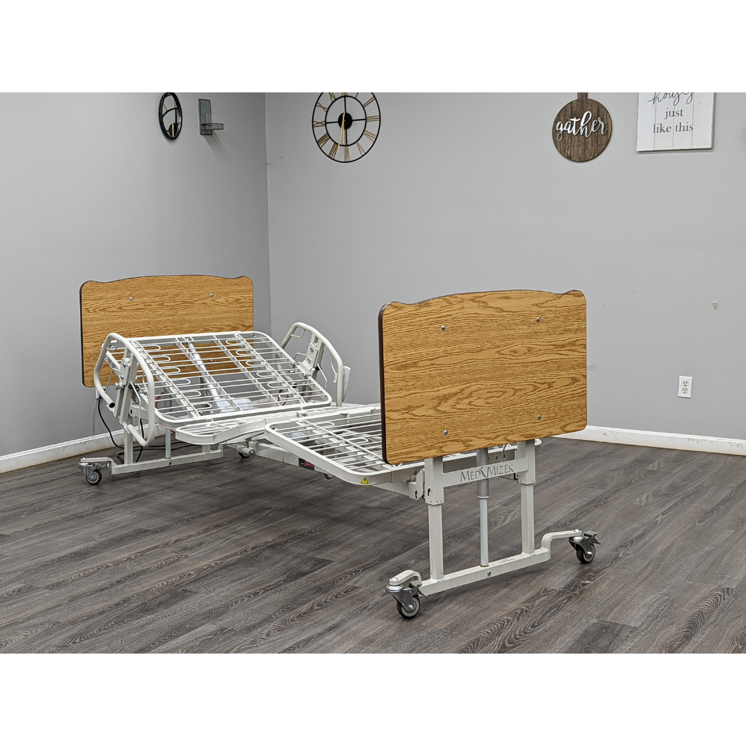 A Med-Mizer AllCare Full Electric Hi/Low Bed - Expandable Width with wooden head and footboards, a metal frame, and wheels is set in a room featuring gray walls, wood flooring, and decorative wall clocks.