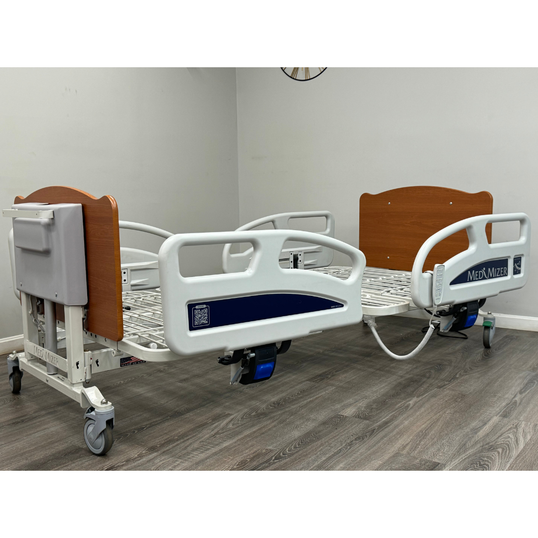 Two Med-Mizer EX8000 ComfortWide Bariatric Width Adjusting Electric Beds with wooden headboards, white metal frames, and side rails sit side by side in a bright room, providing adjustable comfort and support for all patient needs.