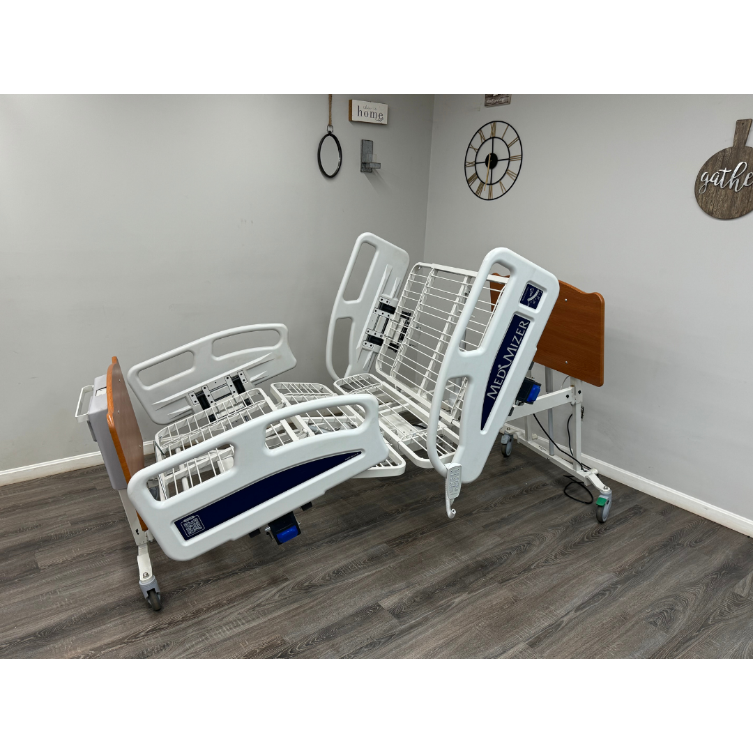 The Med-Mizer EX8000 ComfortWide Bariatric Width Adjusting Electric Bed with metal rails is featured in a modern room with wood flooring, light-colored walls, partially elevated, and wall decorations visible in the background.