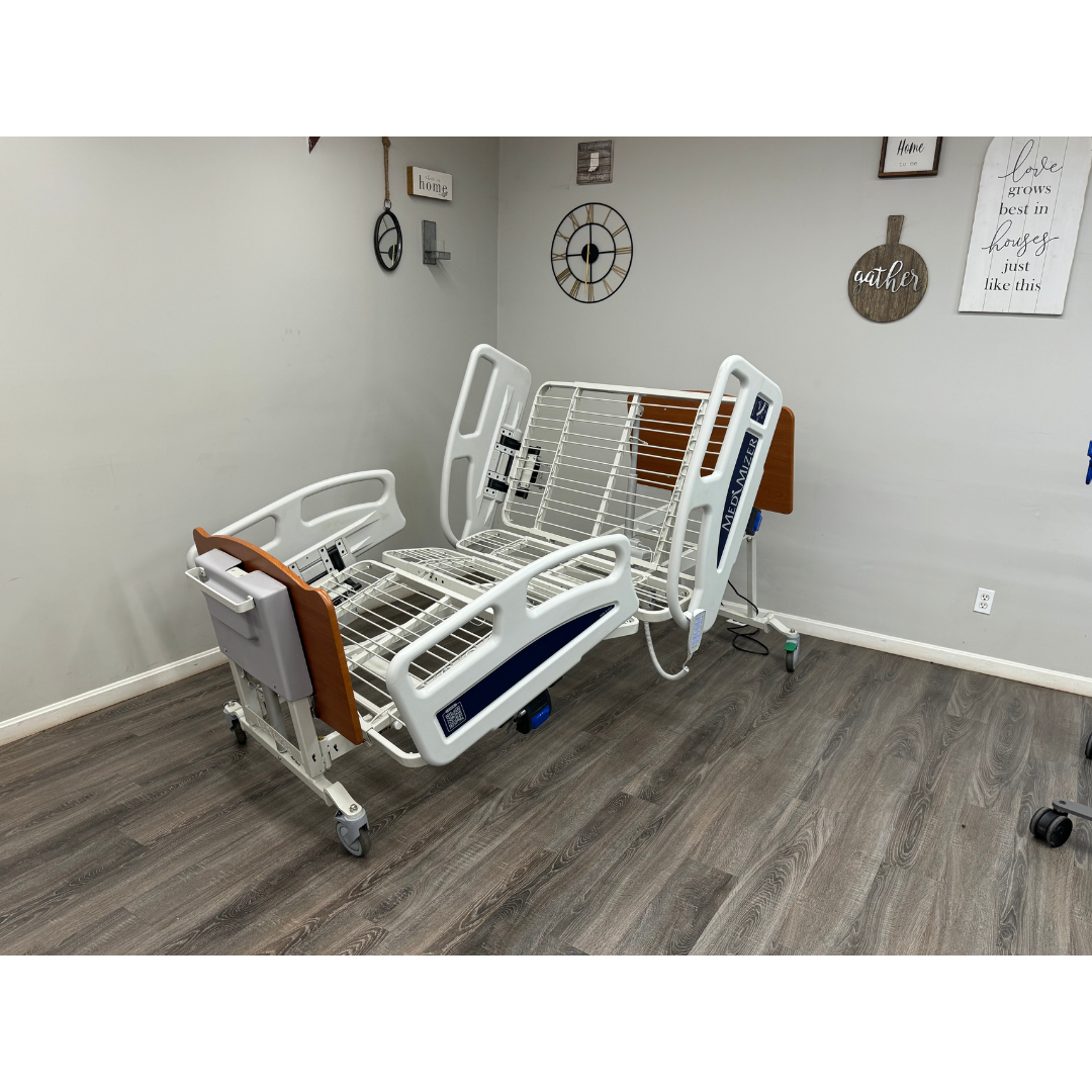 A Med-Mizer EX8000 ComfortWide Bariatric Width Adjusting Electric Bed with raised rails and no mattress stands in a room with wood floors, neutral walls, wall art, and a clock.