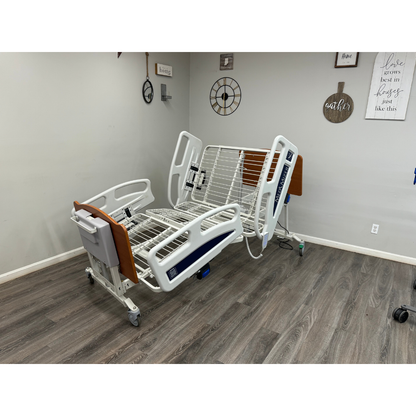 A Med-Mizer EX8000 ComfortWide Bariatric Width Adjusting Electric Bed with raised rails and no mattress stands in a room with wood floors, neutral walls, wall art, and a clock.