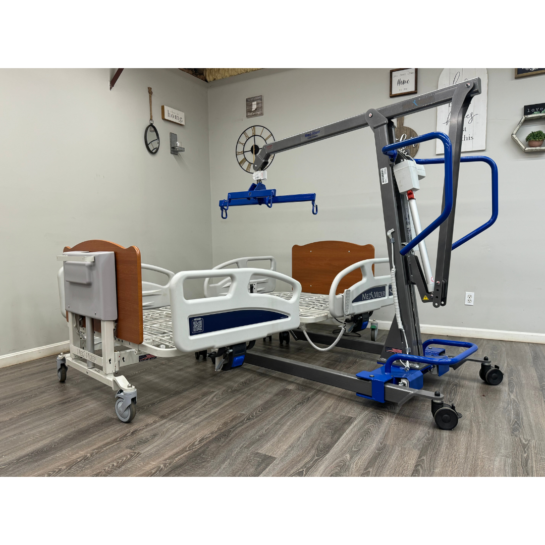 A Med-Mizer EX8000 ComfortWide Bariatric Width Adjusting Electric Bed with side rails and wood-accented headboards sits beside a blue and gray patient lift in a room featuring wood flooring and decorated walls.