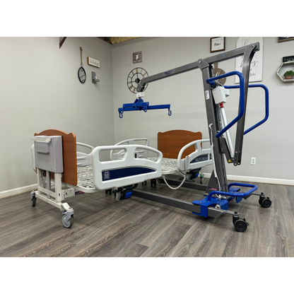 A Med-Mizer EX8000 ComfortWide Bariatric Width Adjusting Electric Bed with side rails and wood-accented headboards sits beside a blue and gray patient lift in a room featuring wood flooring and decorated walls.