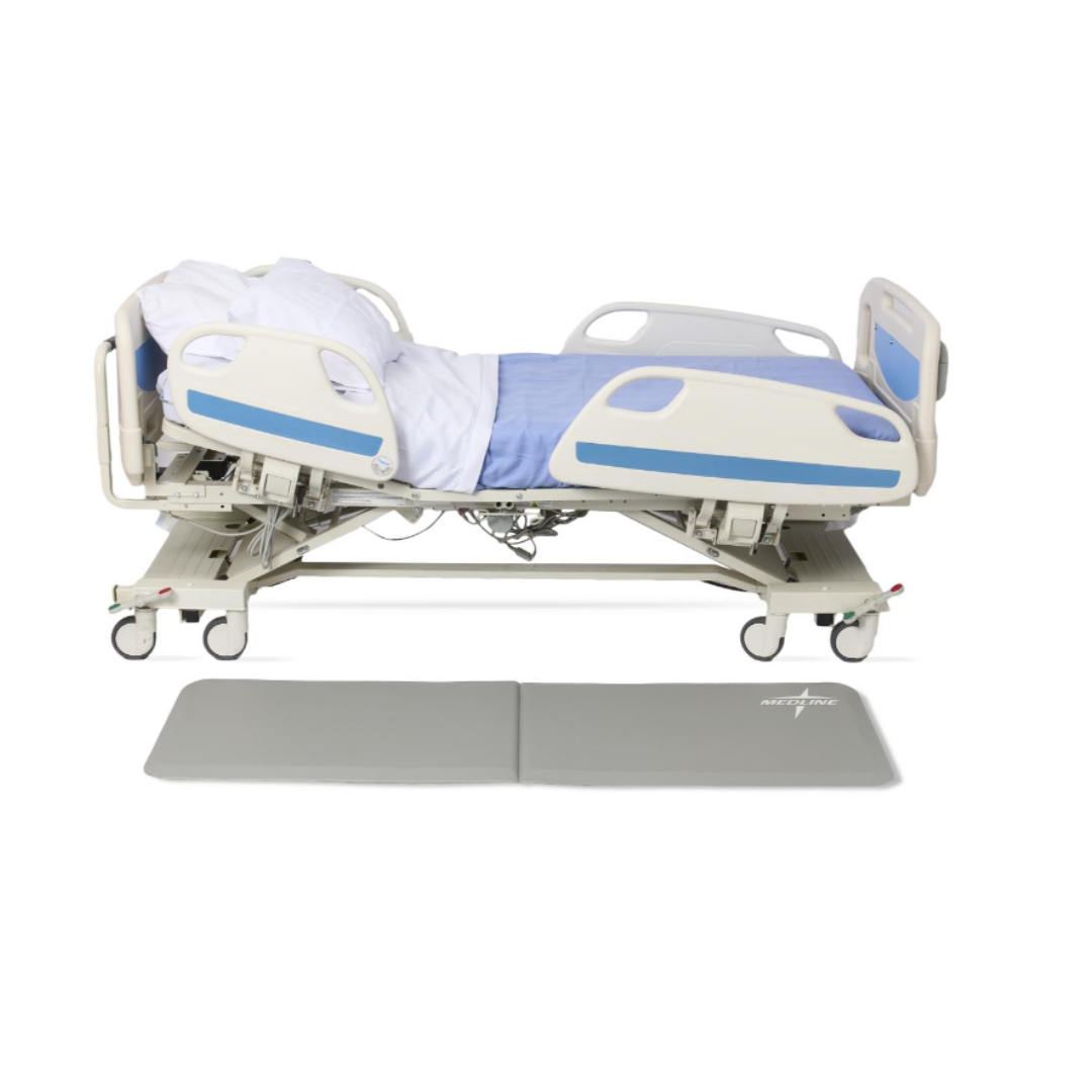 A modern, adjustable hospital bed with white and blue bedding is shown partially raised, with a Medline Bi-Fold Beveled Edge Fall Mat placed beside it on the floor for added safety.