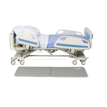 A modern, adjustable hospital bed with white and blue bedding is shown partially raised, with a Medline Bi-Fold Beveled Edge Fall Mat placed beside it on the floor for added safety.